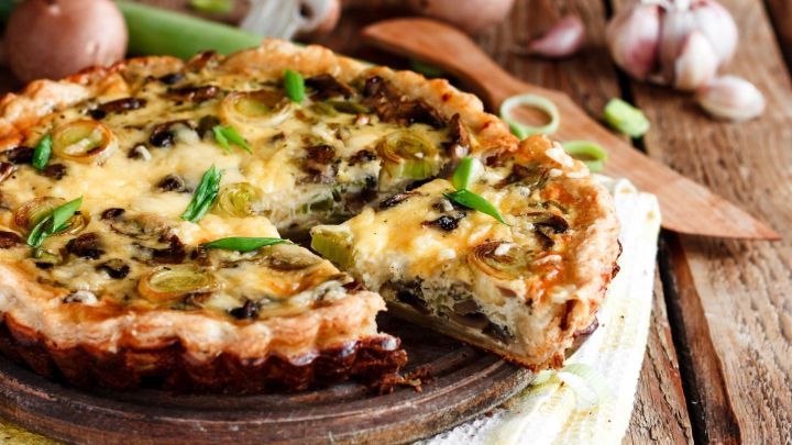 Eine frisch gebackene Champignon-Lauch-Quiche liegt auf einem Holzbrett, umgeben von frischen Zutaten wie Lauch und Knoblauch.