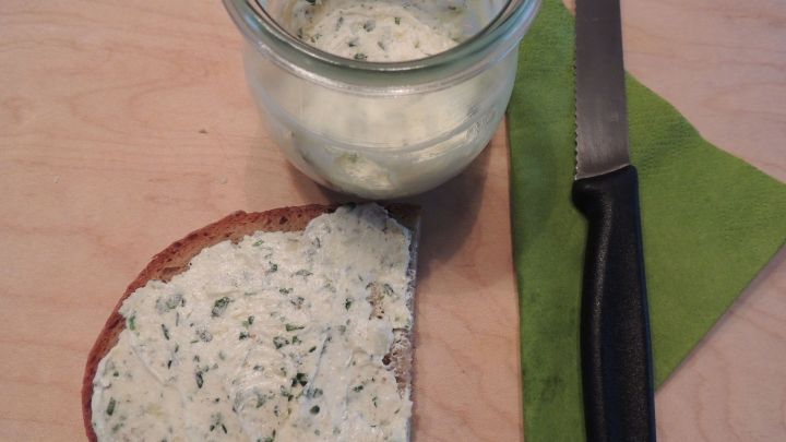 Eine Scheibe Brot ist mit veganer Butter bestrichen, daneben ein Glas mit mehr Butter und ein Messer auf einem Holzuntergrund.