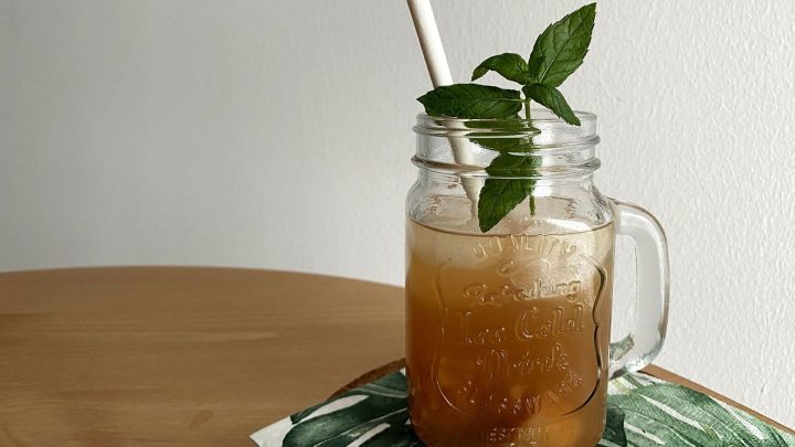 Ein Glas Pfirsich-Eistee Sirup steht auf einem Tisch, dekoriert mit einem Strohhalm und frischer Minze, umgeben von einem Blatt.