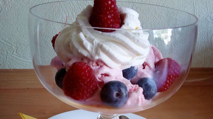 Ein Glas gef&uuml;llt mit Himbeer-Joghurt-Eis, dekoriert mit Sahne und Beeren, auf einem wei&szlig;en Teller mit gelbem Servietten.