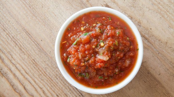 Eine Sch&uuml;ssel mit Knoblauch-Tomaten-So&szlig;e steht auf einem Holztisch, gef&uuml;llt mit einer w&uuml;rzigen, roten Mischung.