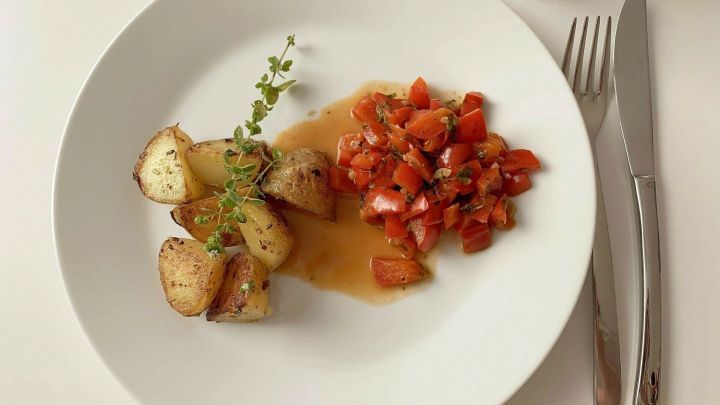 Auf einem wei&szlig;en Teller liegen gebratene Kartoffeln und frisches Paprikagem&uuml;se in einer Sauce, garniert mit Kr&auml;utern.