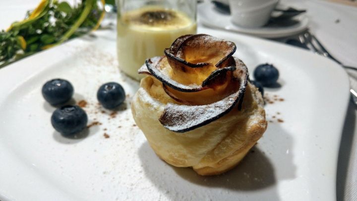 Ein zartes Dessert in Form einer Apfelrose aus Bl&auml;tterteig liegt auf dem Teller, umgeben von Heidelbeeren und Sahne.