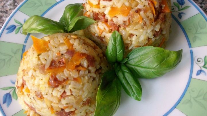 Zwei geformte Reisportionen mit Gem&uuml;se und Salami liegen auf einem bunten Teller, umgeben von frischen Basilikumbl&auml;ttern.