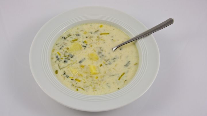 Eine cremige Lauch-K&auml;se-Suppe mit st&uuml;ckigem Gem&uuml;se und einem L&ouml;ffel, serviert in einer wei&szlig;en Sch&uuml;ssel auf hellem Hintergrund.