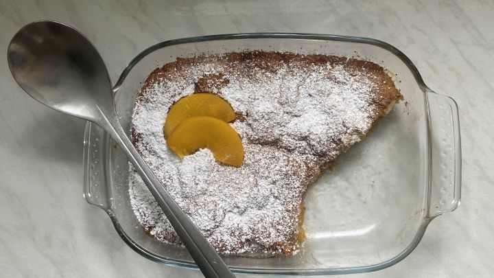 Ein Pfirsich-Cobbler mit Puderzucker und Pfirsichscheiben wird in einer Glasform auf einer Arbeitsfl&auml;che pr&auml;sentiert.