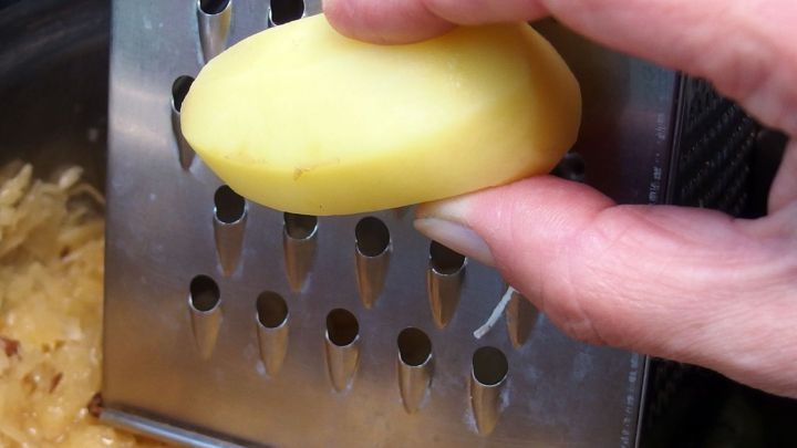 Eine Hand reibt eine Kartoffel auf einer Reibe &uuml;ber einer Sch&uuml;ssel mit Sauerkraut, um das Gericht schmackhafter zu machen.