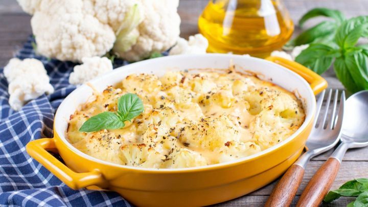 Ein cremiger Blumenkohl-Auflauf mit gr&uuml;nen Basilikumbl&auml;ttern steht bereit, um serviert zu werden, auf einem karierten Tuch.