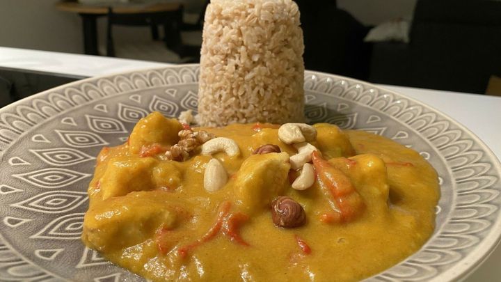 Ein gef&uuml;llter Teller mit Butternuss-K&uuml;rbiscurry und H&uuml;hnchen, umgeben von verschiedenen N&uuml;ssen, serviert mit Reis.