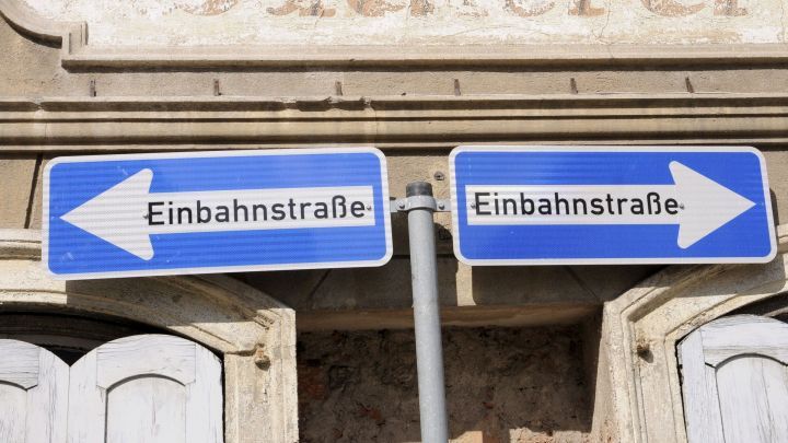 Zwei Verkehrsschilder zeigen die Richtung f&uuml;r "Einbahnstra&szlig;e" nach links und rechts an, vor einem alten Geb&auml;ude.