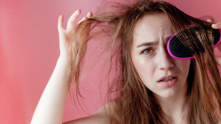 Eine besorgte Frau k&auml;mpft mit ihren fettigen Haaren, w&auml;hrend sie mit einer B&uuml;rste durch ihre Str&auml;hnen geht, vor einem rosa Hintergrund.