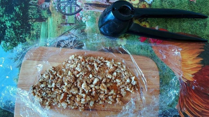 Zerkleinerte N&uuml;sse liegen auf einem Holzbrett, w&auml;hrend ein Nussknacker daneben positioniert ist, auf bunter Tischdecke.