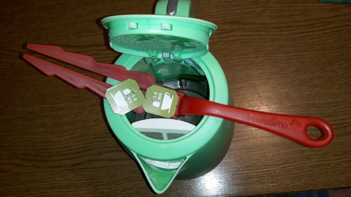 Eine gr&uuml;ne Teekanne steht offen mit roten Zangen, die Teebeutel halten, um ein Herunterfallen beim Tee kochen zu verhindern.