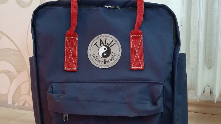 Blauer Rucksack mit roten Griffen steht auf einem Holzfu&szlig;boden vor einer gemusterten Wand und strahlt Handmade-Charakter aus.