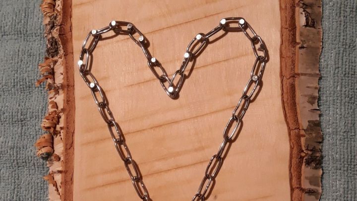 Ein herzf&ouml;rmiges Dekoherz aus Kette auf einem Holzbrett, aufgeh&auml;ngt an einem Faden, vor einem sanften Hintergrund.