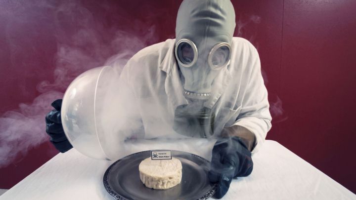 Ein Mensch mit Gasmasken untersucht Stinkek&auml;se, w&auml;hrend Rauch im Raum verbreitet ist, auf einem silbernen Teller.