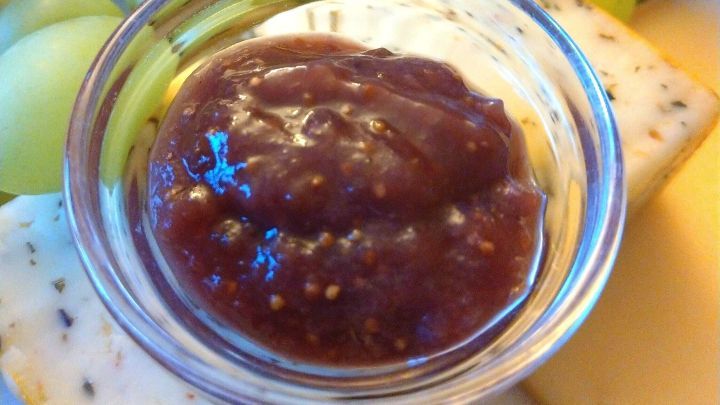 Ein glasierter Becher mit Feigensenf steht zentral, umgeben von K&auml;seplatten und Trauben in einer festlichen Atmosph&auml;re.