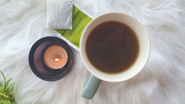 Eine Tasse Brennnesseltee steht auf einem weichen, wei&szlig;en Untergrund neben einer brennenden Kerze und einem Teebeutel.