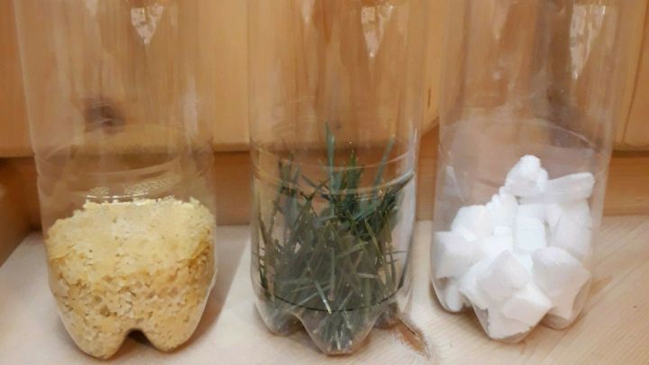 Drei durchsichtige Flaschen stehen auf einem Holzuntergrund, gef&uuml;llt mit verschiedenen Materialien f&uuml;r DIY-Spielzeug.