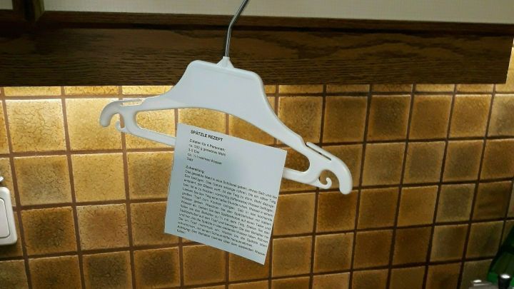Ein Kleiderb&uuml;gel h&auml;ngt an einer Wand, h&auml;lt ein Rezeptpapier; im Vordergrund steht ein Messbecher mit Wasser und einem Nudelholz.