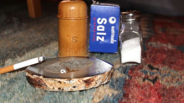 Aschenbecher mit Zigarette und verbranntem Salz befindet sich auf einem bunten Teppich; daneben stehen Salzstreuer und -packung.