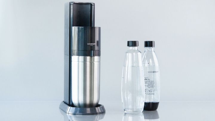 Der SodaStream DUO steht auf einem Tisch, bereit, Wasser in Sprudelwasser umzuwandeln. Daneben zwei Flaschen, eine leer und eine mit Sprudel.