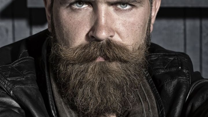 Ein b&auml;rtiger Mann mit intensiven Augen sitzt in einem dunklen Raum und schaut direkt in die Kamera, ausdrucksstark.