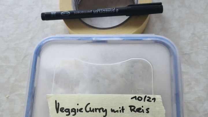Eine durchsichtige Vorratsdose mit &bdquo;VeggieCurry mit Reis&ldquo; beschriftet, umgeben von Malerkrepp und einem schwarzen Marker.