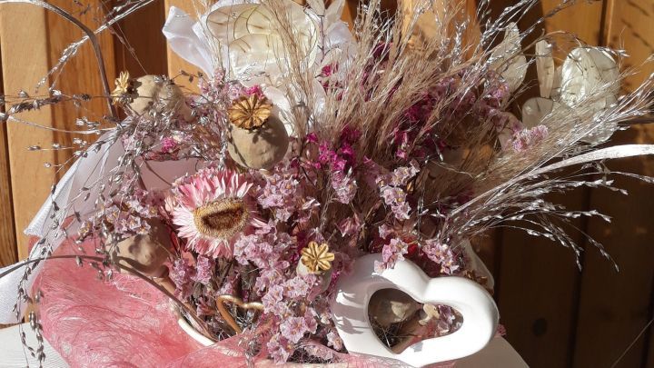 Ein farbenfrohes Herbstgesteck aus Trockenblumen mit rosa Akzenten wird vor einer Holzfl&auml;che pr&auml;sentiert.