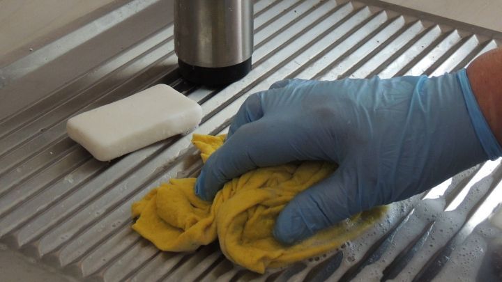 Eine Hand mit blauer Gummihandschuh w&auml;scht mit einem gelben Tuch eine Sp&uuml;le, w&auml;hrend eine Seife daneben liegt.