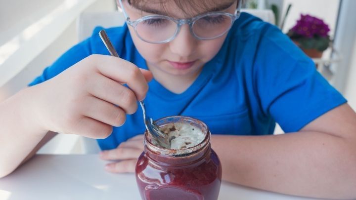 Ein Junge l&ouml;ffelt aus einem Glas Marmelade mit Schimmel, w&auml;hrend er konzentriert am Tisch sitzt.