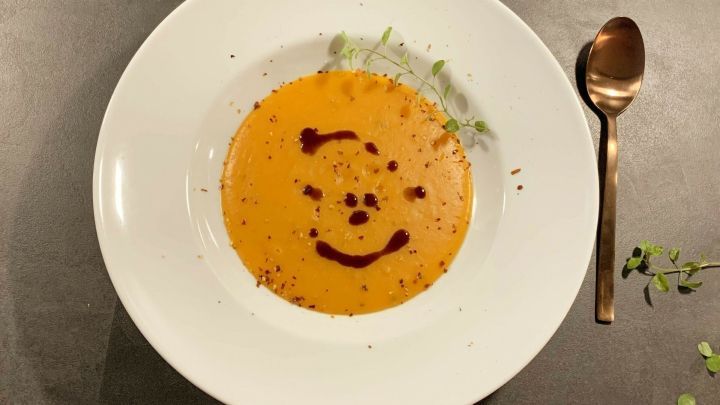 Eine Schale Butternut-K&uuml;rbis-Suppe mit Birne zeigt ein l&auml;chelndes Gesicht aus Sauce und ist von frischen Kr&auml;utern umgeben.