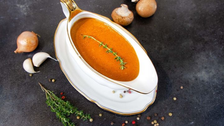 Eine elegante Sch&uuml;ssel mit veganer Bratenso&szlig;e sieht k&ouml;stlich aus und ist umgeben von Zwiebeln, Knoblauch und Pilzen.