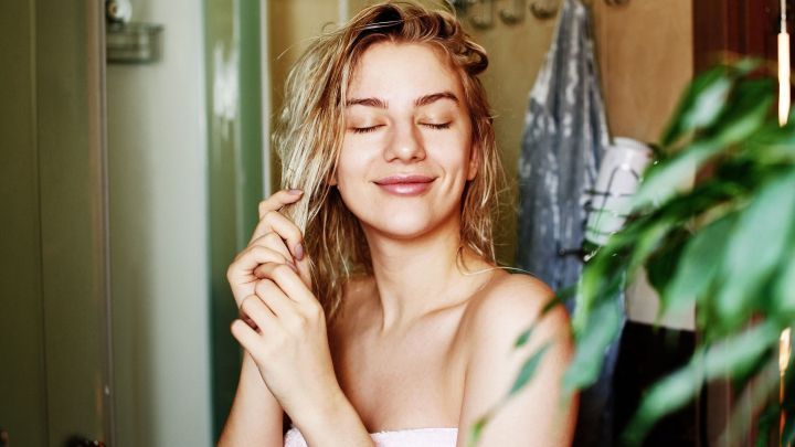 Eine l&auml;chelnde Frau steht in einem Badezimmer und pflegt ihr nasses Haar, umgeben von einer entspannenden, gr&uuml;nen Pflanze.