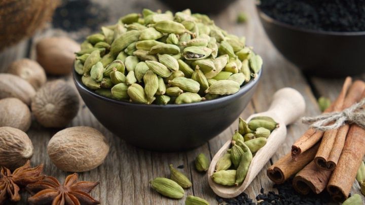 Gr&uuml;ne Kardamomsamen stehen in einer Sch&uuml;ssel, umgeben von anderen Gew&uuml;rzen wie Zimt und Muskatnuss auf einem Holzuntergrund.