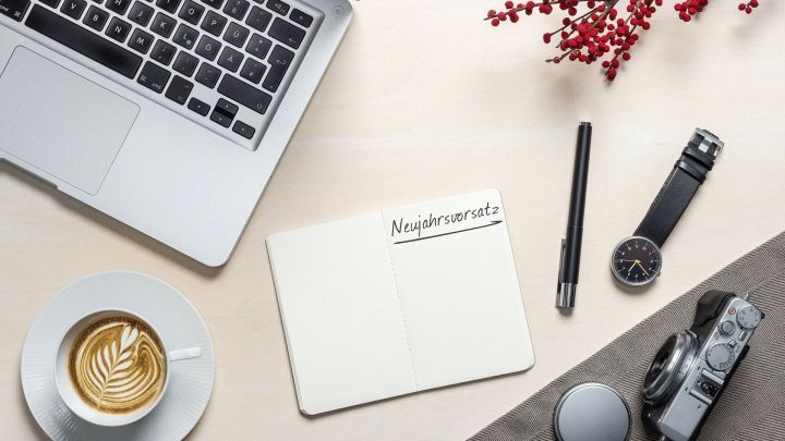 Ein Notizbuch mit dem Wort "Neujahrsvorsatz" liegt neben einem Laptop, einer Tasse Kaffee und weiteren Kreativutensilien.