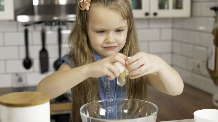 Ein M&auml;dchen bricht vorsichtig ein Ei in eine Sch&uuml;ssel, um R&uuml;hrei zuzubereiten, w&auml;hrend sie in einer hellen K&uuml;che steht.