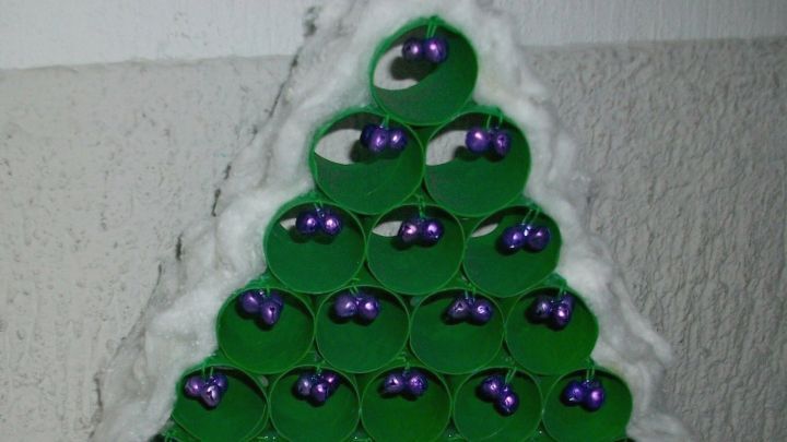 Ein gr&uuml;ner Weihnachtsbaum aus WC-Rollen mit lila Kugeln steht auf einer silbernen Oberfl&auml;che, umgeben von einer Wand.