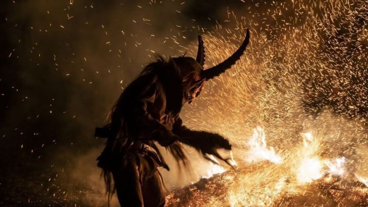 Ein Krampus in einer traditionellen Maske tanzt um ein loderndes Feuer, w&auml;hrend Funken durch die Luft fliegen.
