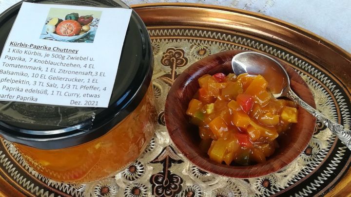 Ein Glas K&uuml;rbis-Paprika-Chutney steht auf einem goldenen Tablett, neben einer Holzschale mit buntem Chutney.