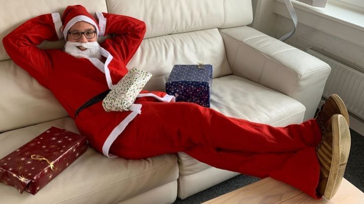 Ein erwachsener Mann in einem roten Weihnachtsmann-Kost&uuml;m entspannt auf einer Couch, umgeben von Geschenken.