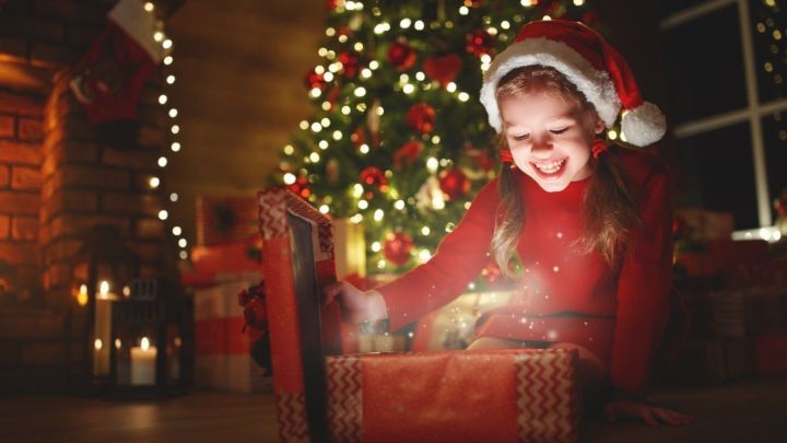 Ein M&auml;dchen in einem Weihnachtsmannkost&uuml;m &ouml;ffnet voller Freude ein glitzerndes Geschenk vor einem festlich geschm&uuml;ckten Weihnachtsbaum.