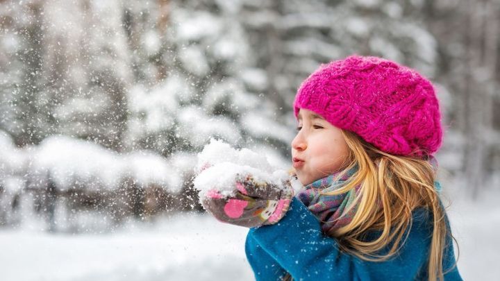 Ein M&auml;dchen mit einem rosa Hut pustet Schnee in der Luft, umgeben von einer winterlichen, verschneiten Landschaft.
