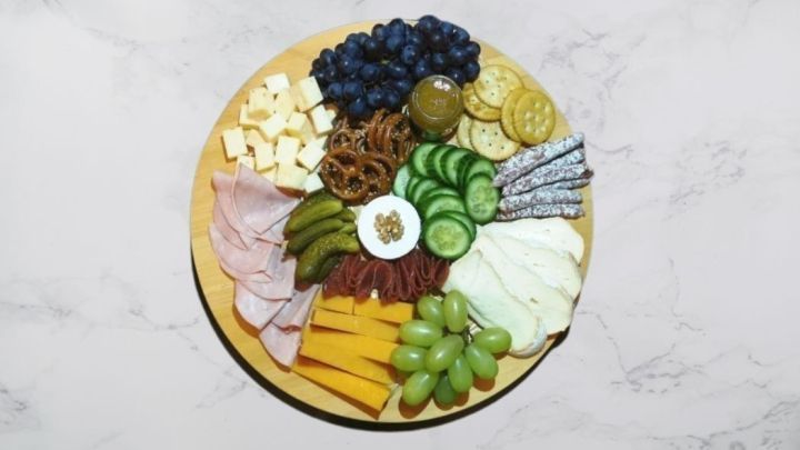 Eine runde Charcuterie-Board zeigt eine Vielzahl von Wurst, K&auml;se, Trauben, Gurken und Crackern, appetitlich angerichtet.