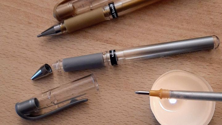 Mehrere Gelroller liegen auf einem Holztisch, w&auml;hrend ein Stift auf einen kleinen, offenen Beh&auml;lter zeigt, um zu schreiben.