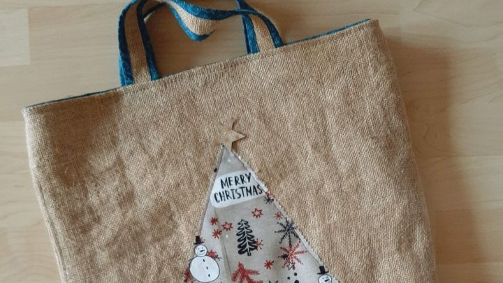 Ein Jutebeutel liegt flach auf einem Tisch und zeigt ein festliches Design mit einem Tannenbaum und Schneem&auml;nnern.