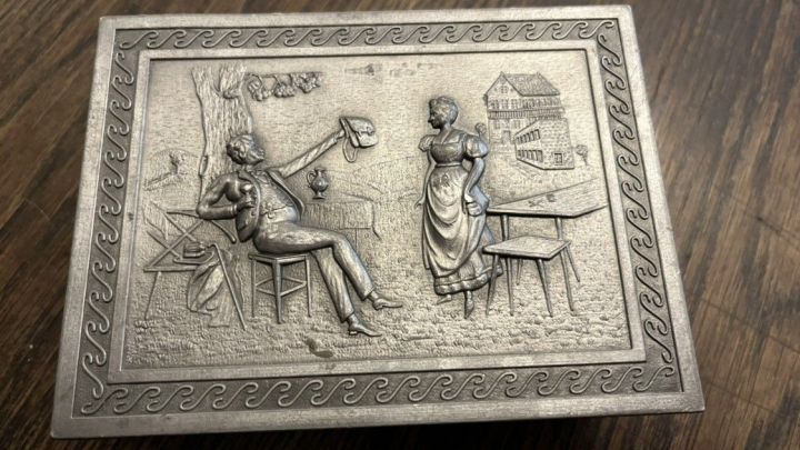 Eine verzierte Zinnbox liegt auf einem Holztisch, w&auml;hrend sie mit Zahnpasta und einer Zahnb&uuml;rste gereinigt wird.