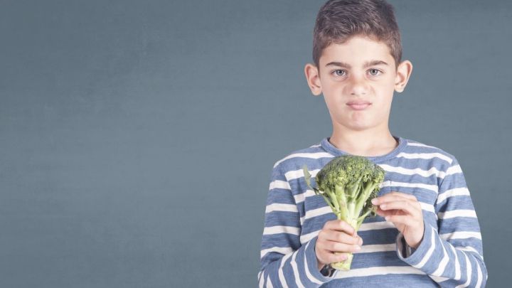 Ein Junge h&auml;lt missmutig einen Brokkoli in der Hand, w&auml;hrend er vor einer grauen Wand steht; gesunde Ern&auml;hrung im Fokus.