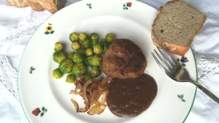 Auf einem Teller liegen gebratene H&uuml;lsenfr&uuml;chte und Pflaumenso&szlig;e, umgeben von Rosenkohl und Brot.