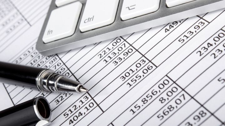 Eine schwarze Kugelschreibermine steht auf einem Finanzblatt, w&auml;hrend eine Tastatur im Hintergrund bereitliegt, um Einnahmen zu verwalten.